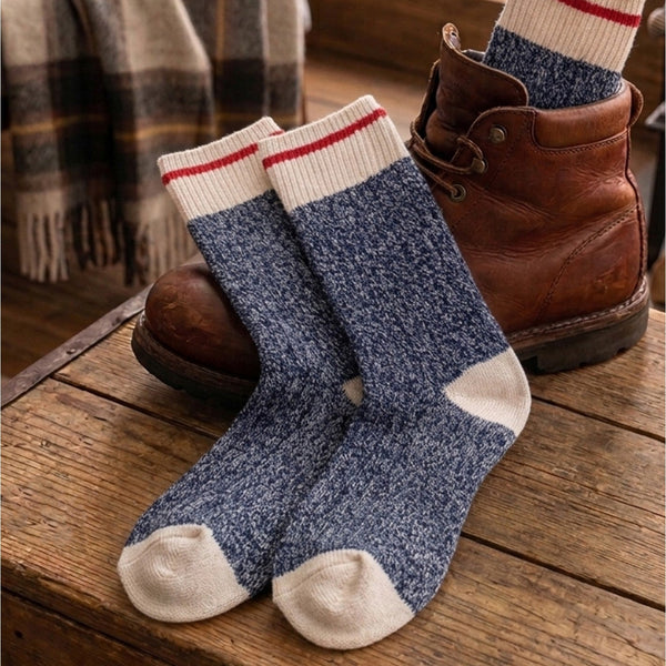A pair of blue marled crew socks with cream toes, heels, and ribbed cuffs with red stripes, styled on a rustic wooden table next to a brown leather boot.