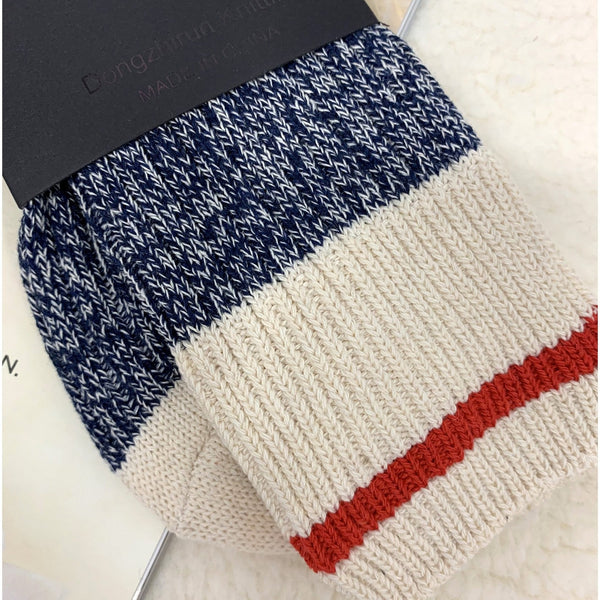 A pair of knit socks folded on a white textured surface, with a dark paper tag attached. The socks have a navy blue and white heathered upper section, a wide cream lower section, and a thin red stripe near the cuff.