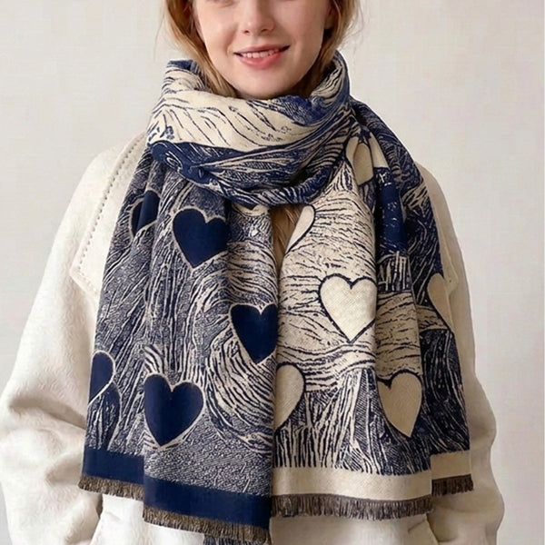 A young woman with blonde hair is wearing a cream and navy blue scarf with a heart and wave pattern, paired with a white coat against a plain light background.