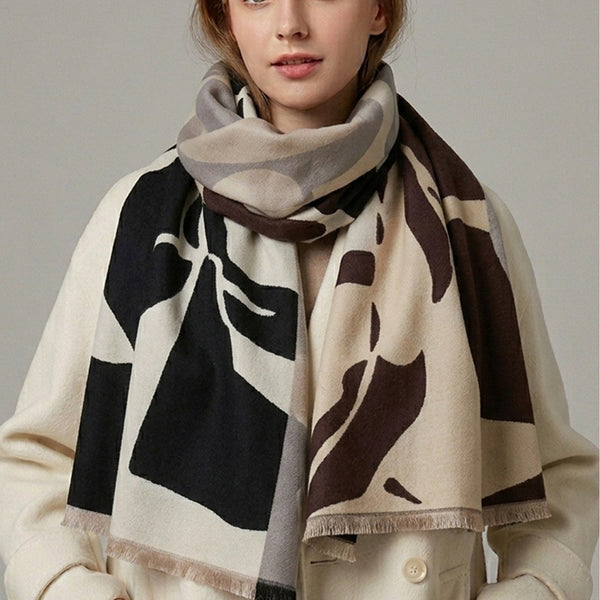A woman wearing a cream-colored coat and a large patterned scarf with abstract black, brown, and gray shapes, photographed against a plain light gray studio background.