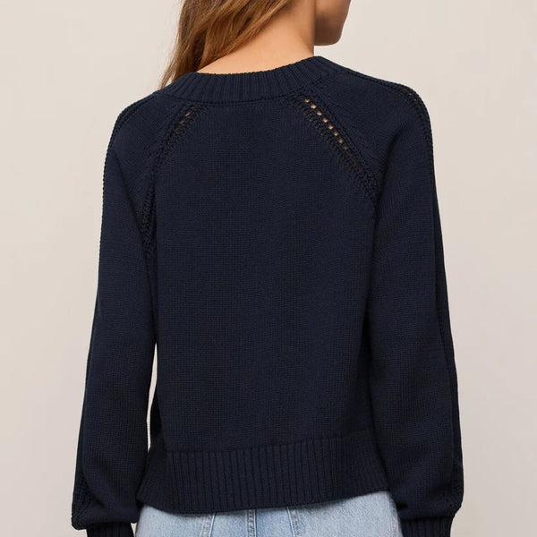 A woman viewed from the back wearing a dark navy knit sweater with decorative openwork details across the shoulders and light blue jeans, against a plain light beige studio background.