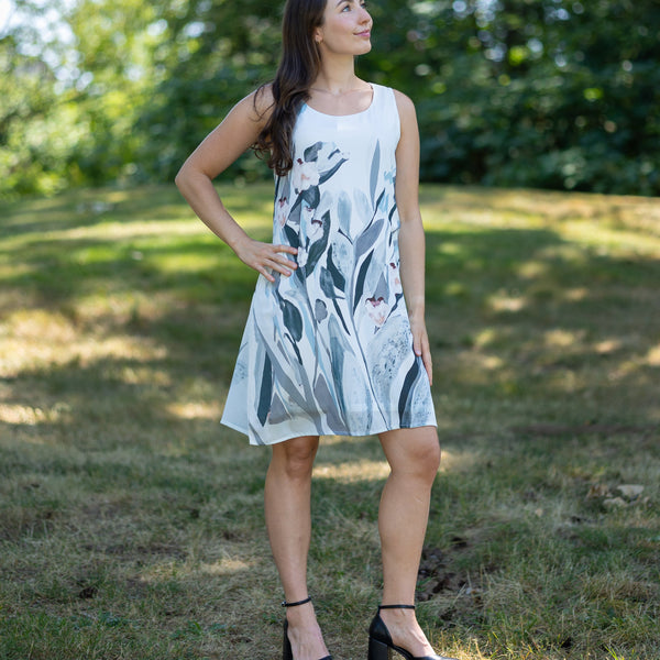 Woman in a white dress with black floral pattern standing in a grassy field with trees in the background