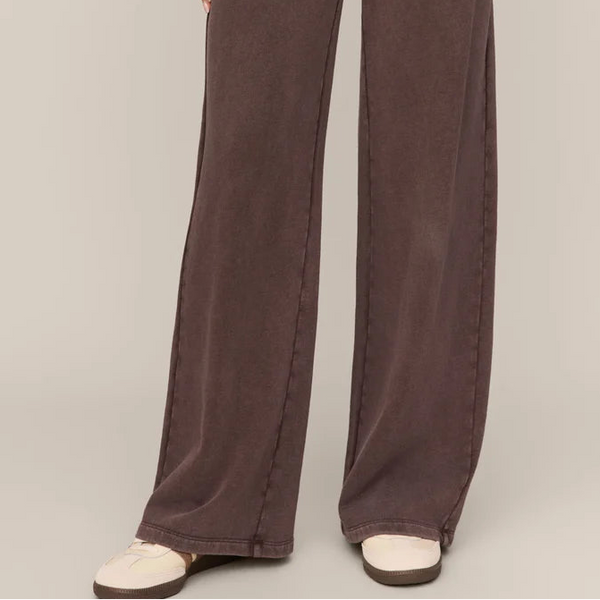 A person wearing wide-leg lounge pants in a washed dark brown or taupe color, paired with a matching cropped sweatshirt and white slip-on shoes, photographed against a plain light gray studio background.