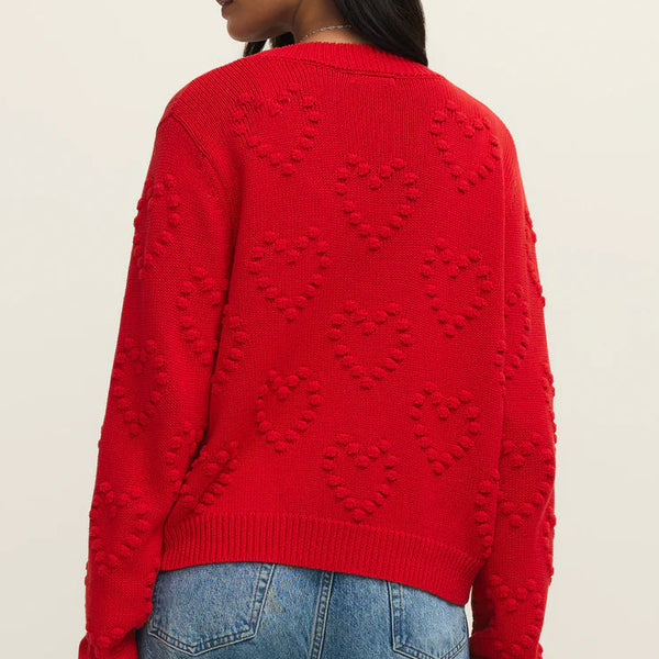 Person wearing a red sweater with heart patterns on the back, standing against a beige background.