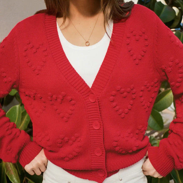 Woman wearing a red knitted cardigan over a white shirt, standing in front of green foliage.