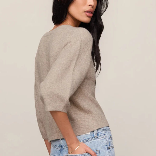 A woman wearing a light taupe knit sweater and light blue jeans, photographed from the back-three-quarter angle against a plain light beige studio background.