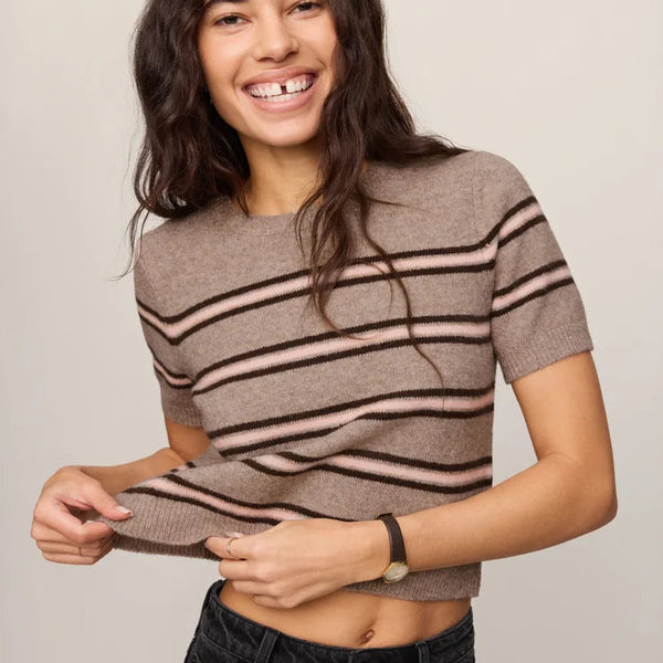 A woman wearing a short-sleeve knit crop top with horizontal taupe and dark brown stripes, paired with black jeans, against a plain light gray studio background.