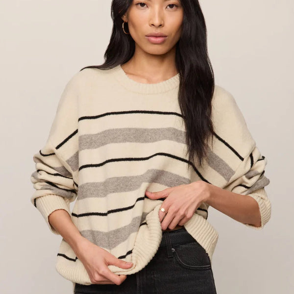 A woman wearing a cream-colored knit sweater with horizontal black and gray stripes, paired with black jeans, against a solid light beige background.