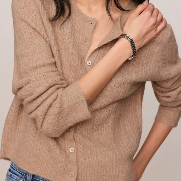 A woman wearing a beige cable-knit button-up cardigan sweater with blue jeans, photographed against a neutral light beige background.