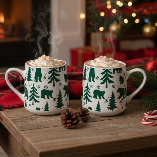 Two white ceramic mugs filled with a hot beverage and topped with whipped cream sit on a wooden table in a cozy holiday setting. The mugs are decorated with a green Christmas tree and bear pattern.