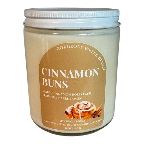 A soy wax candle in a clear glass jar with a white screw-top lid, featuring a tan label that reads 'CINNAMON BUNS' with an image of a cinnamon roll and the phrase 'GORGEOUS WRECK DESIGN'.