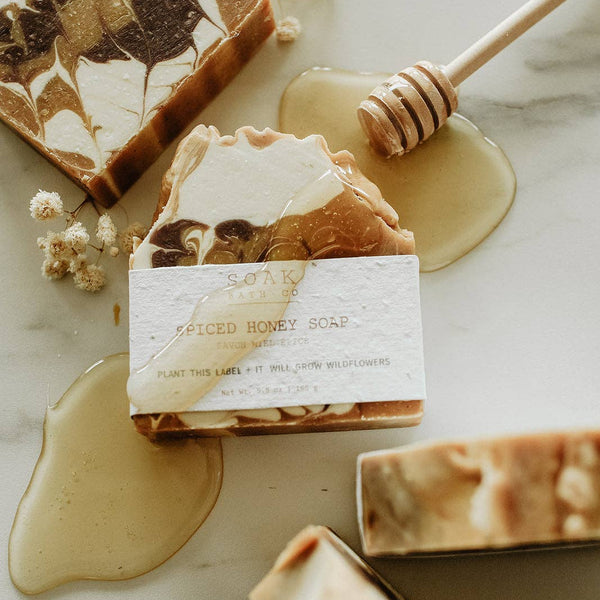 A styled flat-lay of artisanal honey soap bars with a white paper label reading 'SPOON CRAFTED SPICED HONEY SOAP' on a light marble surface, surrounded by honey pools, a wooden honey dipper, and dried white flowers.