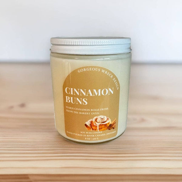 A jar candle labeled 'Cinnamon Buns' by Gorgeous Wreath Designs, sitting on a light wooden surface against a white background.