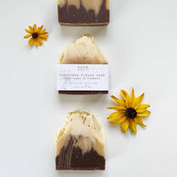 Three handmade soap bars arranged vertically on a white background, with two yellow flower petals placed beside them. The center soap has a paper label reading 'SOAK ARTS CO.