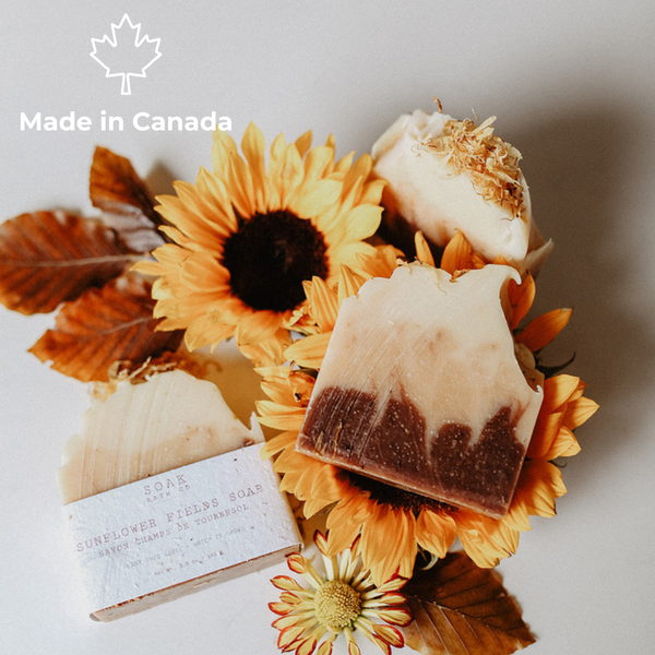 Three handmade soap bars arranged with dried leaves and sunflowers on a light neutral background. One soap bar has a white paper label reading 'SOAP BAR' and 'SUNFLOWER FLESH SOAP'.