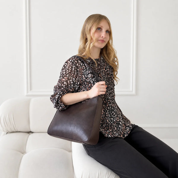 A woman sits on a cream-colored tufted sofa, holding a dark brown leather shoulder bag. She is wearing a brown and beige patterned blouse and black pants, set against a clean white wall with decorative molding.
