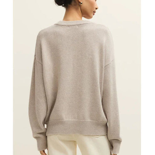 Woman wearing a beige sweater and white pants against a plain background
